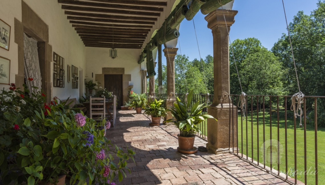 Terraza con arcos y columnas romanicas de la fachada lateral de la masía con vistas al jardín y con la entrada a la capilla al fondo de la imagen 