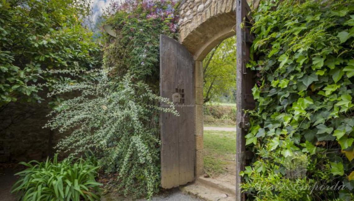Portón de madera de entrada al partió de la masía con el jardín en flor aun lado del portón y una exuberante enredadera en el otro 