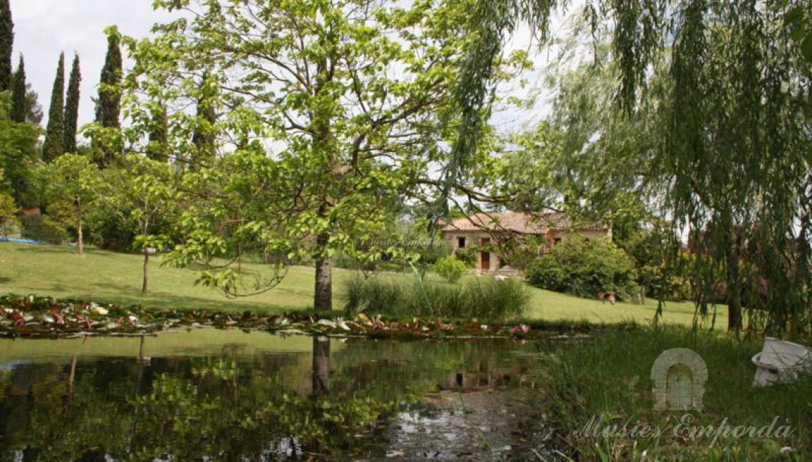 Vista del lago del jardín en primer plano y al fondo la vista de la Masía entre los sauces llorones. 