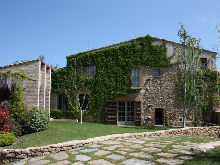 Fachada de piedra de la masía con la yedra trepadora en un lateral y el pabellón de verano con pérgola metálica en el frontal de este    