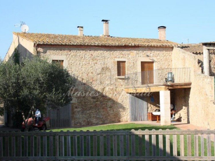 Vista de la fachada de piedra interior de la casa y porche adjunto desde el jardín con valla de madera que protege el acceso a la piscina 